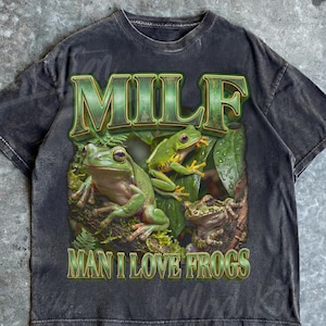 May include: Dark gray t-shirt with a distressed look, featuring a graphic of three green frogs and the text "MILF" and "MAN I LOVE FROGS" in a green, textured font. The shirt has a Pepper color label.