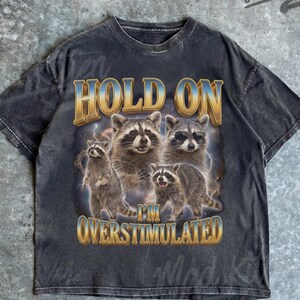 May include: Dark gray t-shirt featuring a graphic of four raccoons with the text "HOLD ON I'M OVERSTIMULATED" in gold and blue. The shirt has a vintage, distressed look and the brand name "Mad Kitten" is visible.