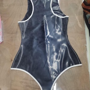 May include: A sleeveless bodysuit made of a dark gray, glossy material with a smooth, reflective finish. The garment features a high-cut leg design and is accented with thin, crisp white trim along the neckline, armholes, and leg openings.