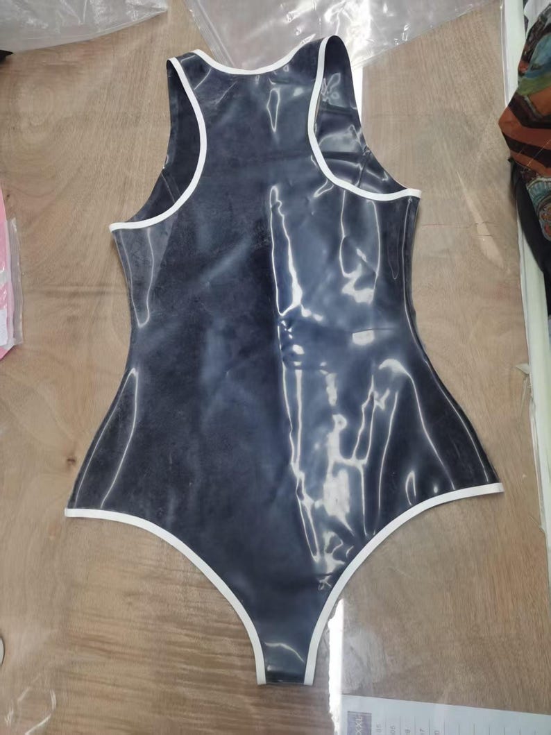 May include: A sleeveless bodysuit made of a dark gray, glossy material with a smooth, reflective finish. The garment features a high-cut leg design and is accented with thin, crisp white trim along the neckline, armholes, and leg openings.