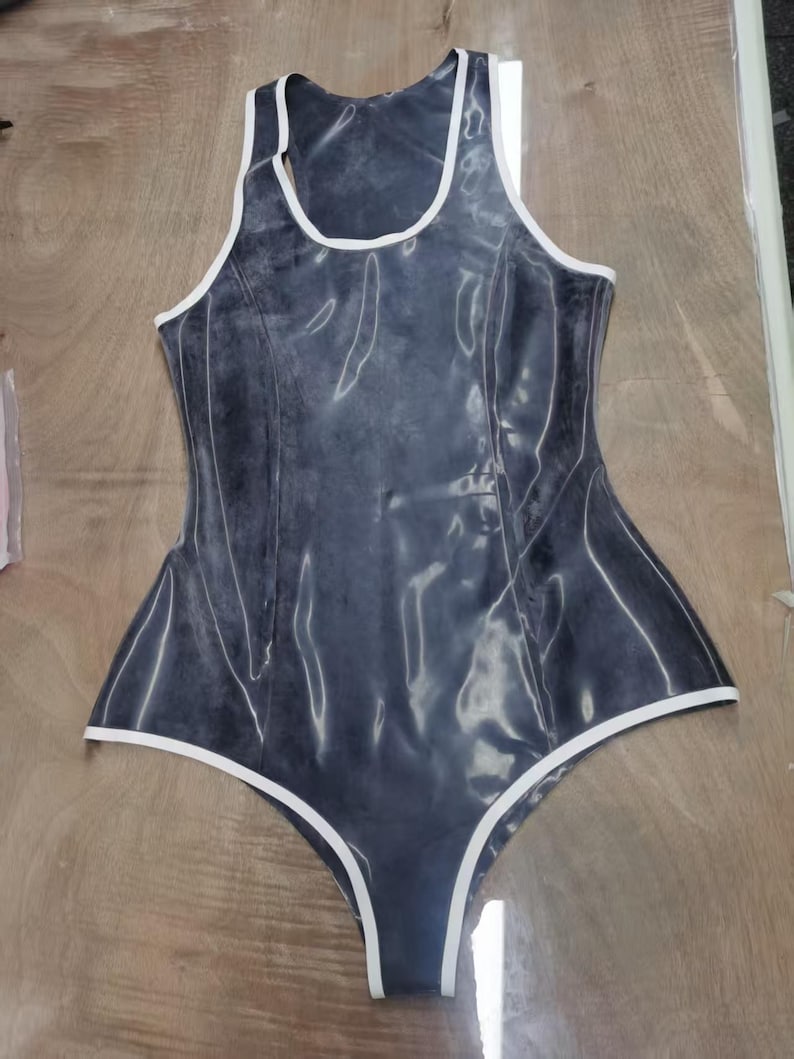 May include: A sleeveless bodysuit in a dark, semi-translucent charcoal gray with a glossy finish. The garment features a high-cut leg design and a scoop neckline, accented throughout with crisp white piping along all the edges and seams.