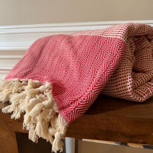 May include: A folded textile throw featuring a vibrant red and white diamond geometric pattern. The fabric is finished with long, cream-colored knotted fringe tassels along the edge, creating a textured appearance on the woven material.