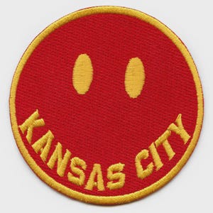 May include: A round, red embroidered patch with a yellow border. The patch features a smiley face with yellow eyes and the words "KANSAS CITY" in yellow, arched along the bottom. The design is simple and bold.