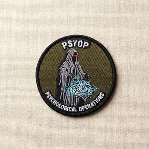 May include: Embroidered patch with a circular design. The patch features a hooded figure with a red glow, holding a blue brain with lightning bolts. The text "PSYOP" is at the top, and "PSYCHOLOGICAL OPERATIONS" is around the bottom edge. The background is olive green.