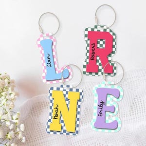 May include: Four personalized letter keychains. The letters are in various colors, including blue, red, yellow, and purple, with names printed on them. Each letter has a checkered pattern border and a silver keyring.