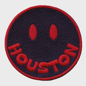 May include: A circular embroidered patch featuring a smiley face design. The patch has a dark blue background with a red border and red eyes. The word "HOUSTON" is embroidered in red across the bottom of the smiley face.
