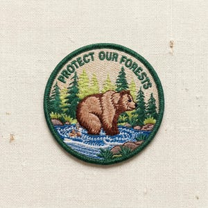 May include: Embroidered patch with a brown bear standing in a river, surrounded by green pine trees. The patch has a green border and the text "PROTECT OUR FORESTS" in a circular design.