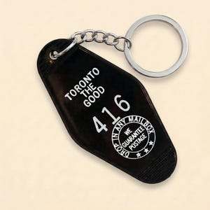 May include: Black motel-style keychain with a silver key ring and chain. The keychain has white text that reads "TORONTO THE GOOD" and "416". A circular graphic states "IN ANY MAILBOX WE GUARANTEE POSTAGE".