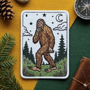 May include: Embroidered patch featuring a brown Bigfoot walking through a forest scene. The patch has a white border and includes trees, stars, a crescent moon, and clouds. The Bigfoot is depicted with a thoughtful expression.