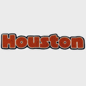May include: A patch with the word "Houston" in orange, textured lettering. The letters are outlined in white and black, with a white background. The patch is likely for clothing or accessories.