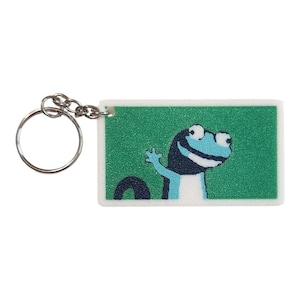 May include: A rectangular keychain featuring a cartoon illustration of a blue and white creature waving from a green background. The keychain has a silver metal ring and chain for attaching keys.