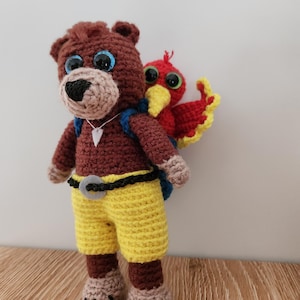 May include: Crocheted bear and bird plush toy. The bear is brown with yellow shorts, a black belt, and blue eyes. The red bird has yellow wings and is attached to the bear's back. The bear has a small white triangle pendant.