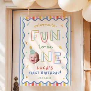 May include: A rectangular birthday sign with a scalloped blue border and colorful striped edges. The text reads "welcome to FUN to be ONE", "LUCA'S FIRST BIRTHDAY!", and "05-09-2026". It features a photo of a baby wearing a blue party hat.