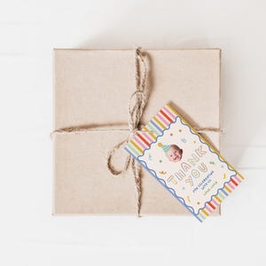 Pu&ograve; includere: Una scatola regalo quadrata in cartone marrone legata con spago di juta naturale, con un'etichetta rettangolare a righe verticali colorate, bordo blu smerlato e il testo THANK YOU FOR CELEBRATING WITH US LOVE, LUCY con foto di un neonato.