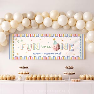Pu&ograve; includere: Uno striscione di compleanno rettangolare con sfondo bianco, bordo a righe colorate e il testo "It's FUN to be ONE" con la foto di un bambino che indossa un cappellino da festa. Lo striscione recita "HAPPY 1ST BIRTHDAY LUCA!" sotto un arco di palloncini.