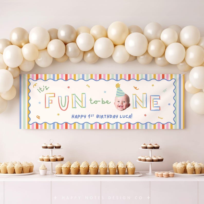 Pu&ograve; includere: Uno striscione di compleanno rettangolare con sfondo bianco, bordo a righe colorate e il testo "It's FUN to be ONE" con la foto di un bambino che indossa un cappellino da festa. Lo striscione recita "HAPPY 1ST BIRTHDAY LUCA!" sotto un arco di palloncini.