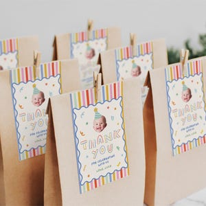 Pu&ograve; includere: Un gruppo di sacchetti di carta marrone, ognuno con un'etichetta rettangolare fissata con una molletta di legno. Le etichette presentano un bordo a righe colorate, la foto di un bambino con un cappellino da festa e il testo "THANK YOU FOR CELEBRATING WITH US LOVE, LUCE".