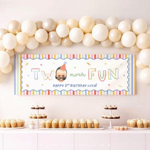 Op de afbeelding: Een rechthoekige verjaardagsbanner met een wit midden, een kleurrijke gestreepte rand en ballonnen erboven. De tekst luidt TWO much FUN en HAPPY 2ND BIRTHDAY LUCA! met een afbeelding van een kind met een feesthoedje en een zonnebril.
