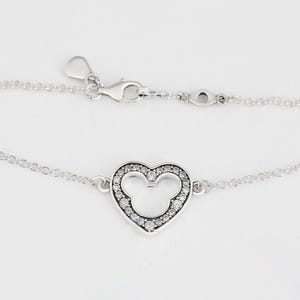 K&ouml;nnte beinhalten: Silbernes Armband mit herzf&ouml;rmigem Anh&auml;nger, der ein Mickey Mouse-Design enth&auml;lt. Der Anh&auml;nger ist mit kleinen, klaren Edelsteinen besetzt. Das Armband hat eine feine Kette und einen Karabinerverschluss.