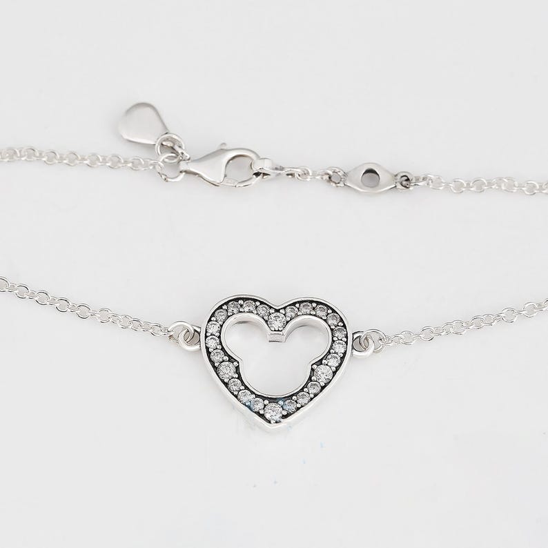 K&ouml;nnte beinhalten: Silbernes Armband mit herzf&ouml;rmigem Anh&auml;nger, der ein Mickey Mouse-Design enth&auml;lt. Der Anh&auml;nger ist mit kleinen, klaren Edelsteinen besetzt. Das Armband hat eine feine Kette und einen Karabinerverschluss.
