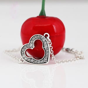 K&ouml;nnte beinhalten: Ein silbernes Armband mit einem herzf&ouml;rmigen Anh&auml;nger, besetzt mit klaren Kristallen. Der Anh&auml;nger hat ein von Mickey Mouse inspiriertes Design. Eine gl&auml;nzende rote Kirsche ist im Hintergrund. Das Armband liegt auf einer reflektierenden Oberfl&auml;che.