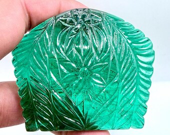 May include: A carved, emerald-green gemstone with a floral and leaf design. The gemstone is held in a hand and features intricate details, including a central flower and surrounding foliage. The stone has a polished, reflective surface.