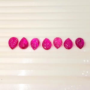 Puede incluir: Siete gemas fucsia en forma de hoja están dispuestas horizontalmente. Cada gema tiene vetas detalladas talladas en su superficie, dando una apariencia realista. Las gemas se colocan sobre un fondo blanco y acanalado, creando un contraste.
