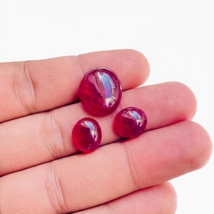 Op de afbeelding: Drie gepolijste, ovaalvormige, dieprode edelstenen. De edelstenen variëren in grootte, met één grotere steen en twee kleinere. De stenen zijn glad en reflecteren licht, wat wijst op een hoge glans.