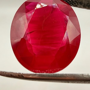 May include: A faceted, oval-shaped ruby gemstone, held by a pair of silver tweezers. The ruby is a deep, saturated red color, with visible internal inclusions and a high degree of transparency. The gemstone is polished and reflects light.
