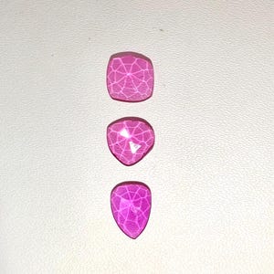 Op de afbeelding: Drie roze edelstenen in verschillende vormen: een vierkant, een afgeronde driehoek en een druppel. Elke edelsteen heeft een webachtig patroon over het oppervlak, wat een textuur geeft. De edelstenen zijn verticaal geplaatst tegen een witte achtergrond.
