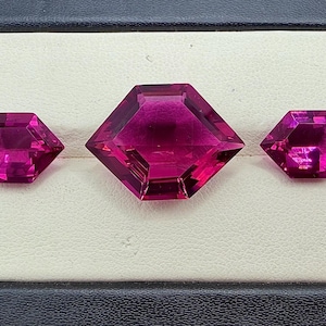 May include: Three faceted, hexagonal gemstones in a deep magenta hue are displayed in a black jewelry box. The central gem is larger than the two flanking it. The gems are set against a white background.