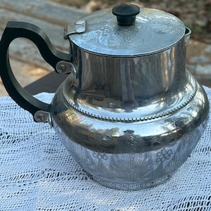 Könnte beinhalten: Eine silberfarbene Vintage-Teekanne mit schwarzem Griff und Deckelknopf. Die Teekanne hat ein geprägtes Blumendesign am Körper und am Deckel. Der Griff ist gebogen und mit silbernen Nieten befestigt. Die Teekanne steht auf einer weißen, strukturierten Oberfläche.