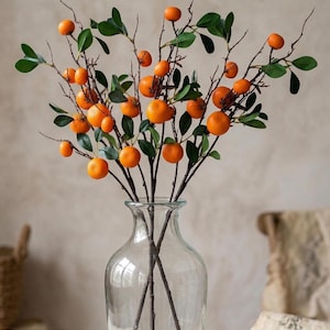 May include: A clear glass vase holds branches with vibrant orange fruit and green leaves. The branches are arranged to create a natural, decorative display. The vase sits on a wooden surface, suggesting a home decor theme.
