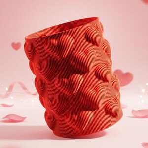 Può includere: Un contenitore cilindrico rosso con un motivo a forma di cuore. Il contenitore ha una texture a coste ed è circondato da cuori rosa e petali di rosa. Il design è moderno e romantico, adatto per San Valentino o altre occasioni speciali.