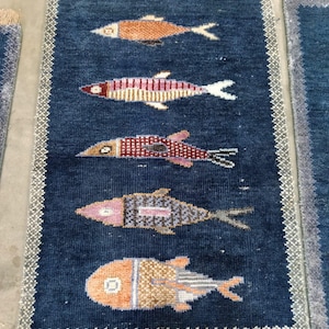 May include: A rectangular dark blue rug featuring five stylised fish motifs arranged vertically. Each fish displays a unique pattern and colour combination, including orange, red, and purple tones. The rug has a light grey border and tan fringe.