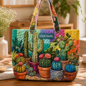 Puede incluir: Un bolso tote acolchado con un colorido diseño de jardín de cactus. El bolso tiene forma rectangular con un asa estampada floral y el texto "YOUR NAME" en una etiqueta azul. El bolso muestra varios cactus en macetas de terracota.