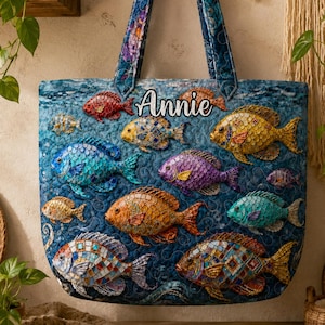 May include: A quilted tote bag with a blue background and colourful fish designs. The bag features a variety of fish in shades of blue, orange, yellow, and purple. The name "Annie" is embroidered on the bag. The bag has blue handles.