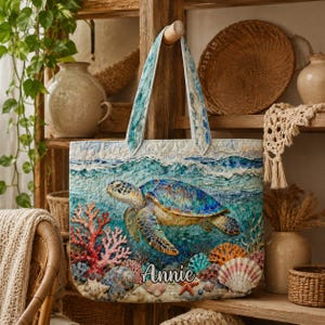 May include: A quilted tote bag with a sea turtle design, coral, and seashells. The bag has blue and white handles and the name "Annie" is printed on the front. The bag is displayed against a rustic wooden shelf. The bag's design evokes a marine theme.
