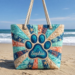 May include: A quilted tote bag with a beach theme, featuring a large blue paw print with the name "Sophia." The bag has rope handles and is decorated with seashells, starfish, and coral in shades of blue, orange, and beige.