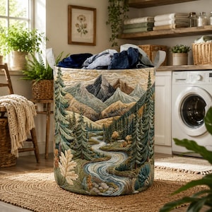 Pode incluir: Um cesto de roupa cilíndrico de tecido com um design acolchoado de paisagem que mostra um rio sinuoso, densas florestas de coníferas e montanhas imponentes. O exterior apresenta costuras detalhadas em tons de verde, azul e bege com duas alças.