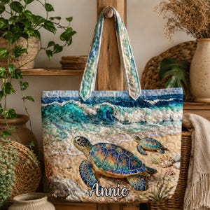 May include: A quilted tote bag depicting a beach scene with turquoise waves and two sea turtles on the sand. The bag has blue and green handles and the name "Annie" is printed on the front.
