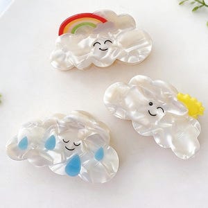 Op de afbeelding: Drie haarclips in de vorm van een wolk met lachende gezichten. Eén clip heeft een regenboog, een andere blauwe regendruppels en de derde een gele zon. De clips zijn wit met een parelmoer afwerking.