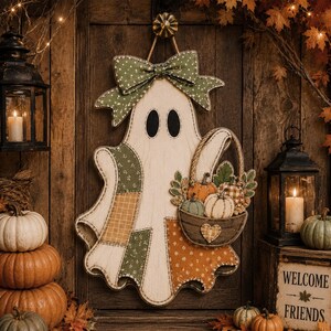 May include: A decorative wooden ghost wall hanging featuring a green polka dot bow, patchwork fabric patterns in green, orange, and yellow, and a basket filled with small pumpkins. The ghost has two black oval eyes and a small heart on the basket.