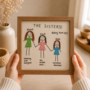 May include: A square wooden frame containing an embroidered artwork titled THE SISTERS! featuring three stick figures named Nicole, Ellie, and Auza holding hands. Each figure wears a colored dress and has descriptive text embroidered below them.