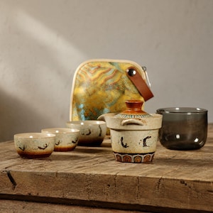 May include: A ceramic tea set with a teapot, four small cups, and a dark glass cup. The teapot and cups have a beige and brown color scheme with decorative patterns. A yellow and blue patterned carrying case is in the background.