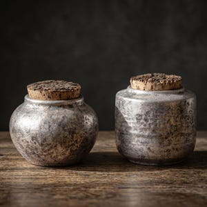 May include: Two metallic containers with a distressed silver finish and textured surfaces. One container has a rounded, bulbous shape, while the other is cylindrical. Both vessels are topped with natural, porous cork stoppers.