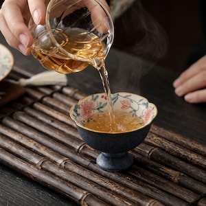 Puede incluir: Una jarra de cristal transparente con diseño acanalado vierte té ámbar en un pequeño cuenco de cerámica con pie. El cuenco presenta una base azul oscuro moteada y un borde interior blanco decorado con un delicado patrón floral rosa y hojas verdes.