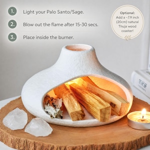 May include: A white ceramic burner with an opening to hold Palo Santo sticks. The burner sits on a wooden coaster, with two clear quartz crystals and a lit candle nearby. Instructions are printed on the image.
