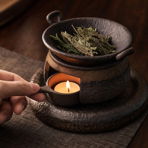 May include: A dark brown, textured ceramic oil burner with a wide bowl containing dried green herbs. The base features a circular opening holding a small, lit tea light candle in a matching ceramic holder with a handle, all set on a round tray.