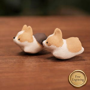 May include: Two small, ceramic corgi figurines. The corgis are white, tan, and gray, with black eyes and noses. The figurines are lying down on a wooden surface. A gold seal with the words "Free Engraving" is in the bottom right corner.